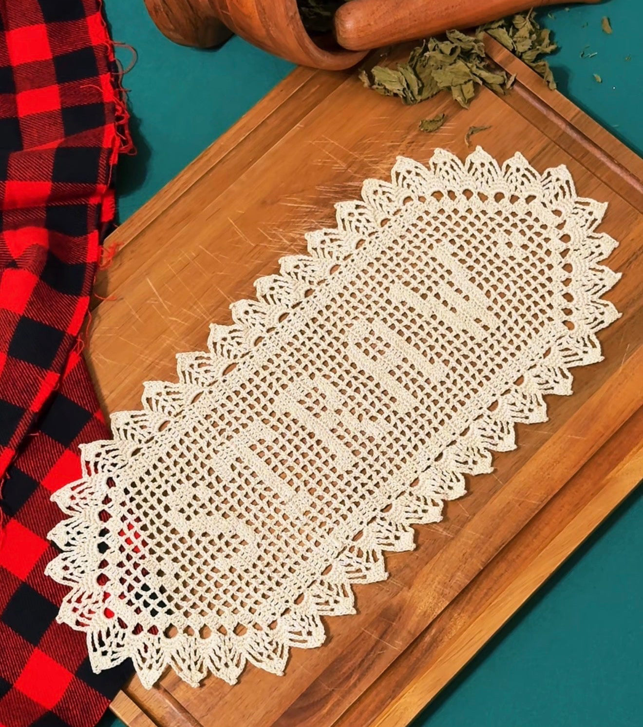 An off-white/tan colored handmade, custom order name doily displayed on a wooden, rustic cutting board. The background contains a red/black checkered flannel. 
